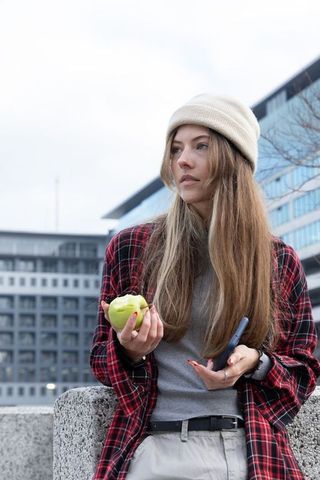 Young woman leaning on concrete ledge holding apple and phone in urban casual streetwear