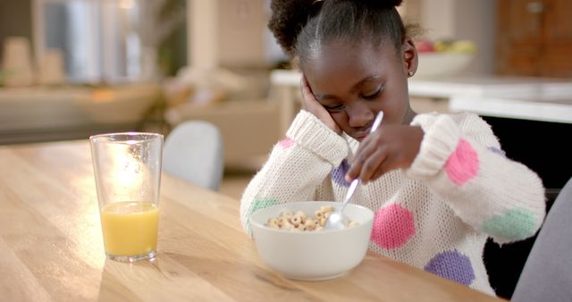 Young girl is sleepily eating a bowl of cereal with a glass of orange juice at a wooden table. The scene suggests a typical morning routine in a comfortable home kitchen. Ideal for articles on children's morning routines, nutrition, or parenting tips.