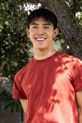 Smiling young man leaning against tree in rust red t-shirt under dappled sunlight