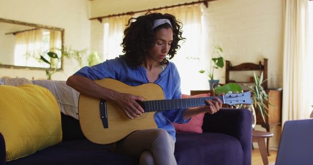 Biracial woman playing guitar while sitting on the couch in front of laptop at home. staying at home in self isolation in quarantine lockdown