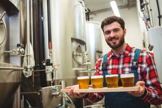 Brewery worker holding a tray with beer samples in an industrial brewing environment. Ideal for use in articles or advertisements related to craft beer, brewing processes, quality control in beverage production, or the craft beer industry. Can also be used in promotional materials for breweries or beer tasting events.
