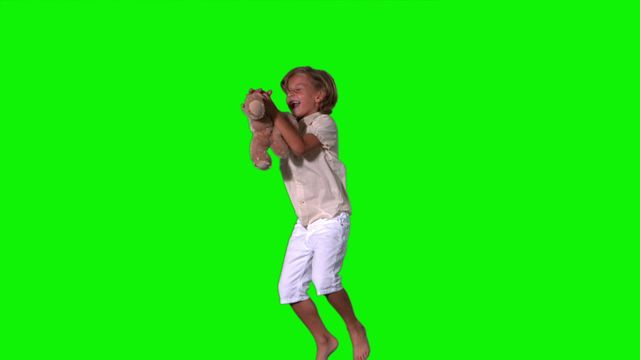 Boy jumps while holding a teddy bear against a green screen. He appears very excited and playful, capturing a sense of joy and energy. Ideal for use in advertisements, educational content, and promotions involving children, toys, or happiness.