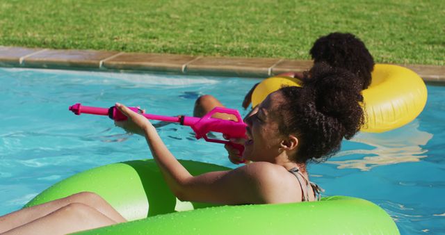 Young girls having fun by playing with water guns in a pool on a sunny summer day. Ideal for themes of childhood joy, outdoor activities, summer fun, and leisure. Great for use in advertisements, vacation brochures, and social media posts promoting pool toys and summer activities.