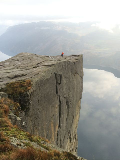 Person standing alone on the edge of a tall, sheer cliff overlooking fjords in Norway. The surrounding landscape is dramatic with rolling mountains and calm waters. Useful for travel blogs, adventure themes, and promoting tourism and outdoor activities in Norway.