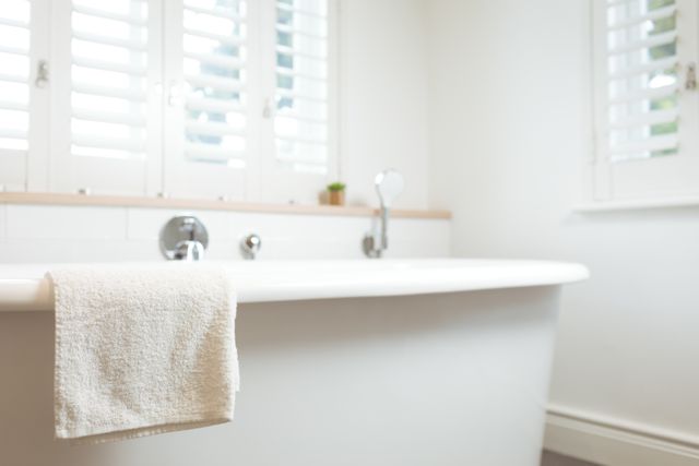 This image showcases a modern bathroom featuring a white bathtub with a towel draped over the edge. The room is bright and clean, with natural light streaming through the shutters. Ideal for use in home decor magazines, interior design blogs, and advertisements for bathroom products or cleaning services.