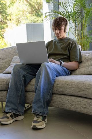 Teen using laptop on sofa with headphones and smartwatch in modern minimal home interior