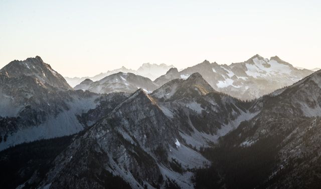 Photograph of a majestic mountain range illuminated by the soft light of sunrise. Snow-capped peaks stand in contrast to the darker, rocky terrain, creating a stunning visual landscape. Ideal for use in travel brochures, nature blogs, inspirational posters, or to promote outdoor adventure activities.