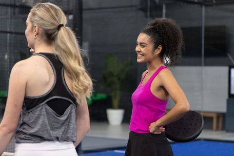 Smiling diverse women on indoor padel court holding paddles and sharing laugh during warmup
