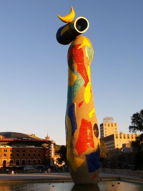 The image features a Joan Miró sculpture with vivid colors in an urban park during sunset. This photo can be used for travel guides, art articles, or city brochures to showcase recognizable landmarks and cultural attractions.