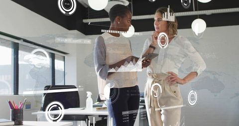 Female colleagues collaborating with tablet and digital dashboards in sunlit modern office