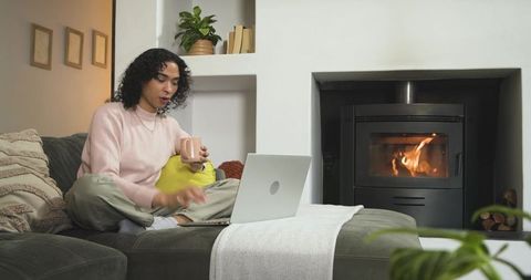 Non-binary person relaxing with laptop and mug on sofa by fireplace in cozy living room