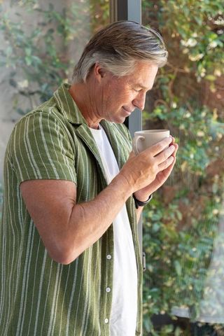 Mature man enjoying morning coffee by sunlit window with lush greenery, relaxed casual vibe