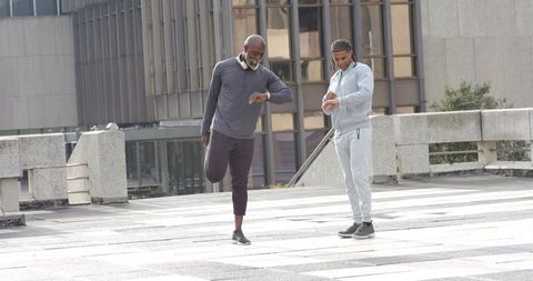Diverse men stretching and checking fitness trackers on urban rooftop plaza