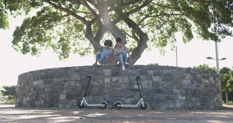 Two Women Relaxing Outdoors With Electric Scooters Under Tree
