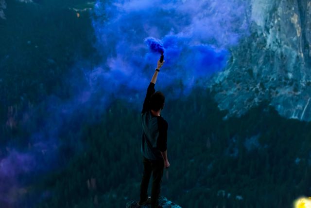 Man standing at the edge of a mountain, holding a purple smoke bomb high in the air. Scenic and adventurous setting with misty blue and purple smoke floating around. Perfect for adding drama to travel blogs, adventure branding, and inspirational posters.