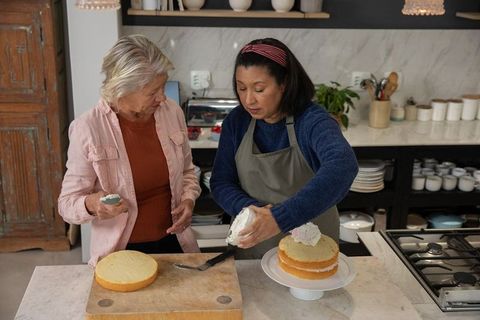 Mature women frosting layered sponge cake in cozy kitchen, baking together and sharing skills