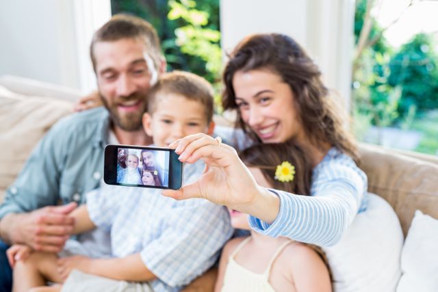 Family sitting on a couch, smiling and taking a selfie with a mobile phone. Perfect for use in advertisements, family-oriented content, technology promotions, and lifestyle blogs.