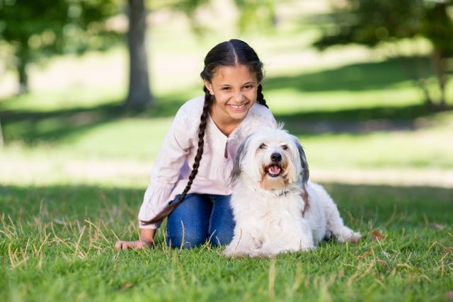 Young girl with long braids enjoying time with her fluffy white dog in a sunny park. Ideal for themes of childhood joy, pet companionship, outdoor activities, and family leisure. Perfect for advertisements, blogs, and articles about pets, children, and outdoor fun.