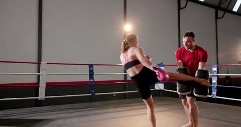 Female fighter delivering high kick to male trainer holding pads inside boxing ring