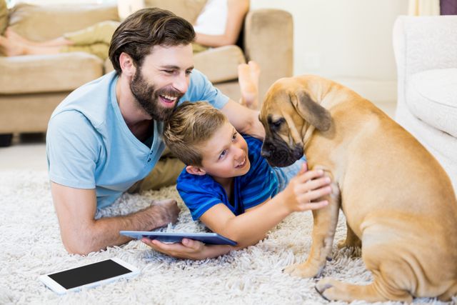 Father and son lying on carpet in living room, bonding with their dog while using a digital tablet. Ideal for themes of family bonding, technology in daily life, pet companionship, and modern parenting. Suitable for advertisements, blogs, and articles about family life, pet care, and digital interaction.
