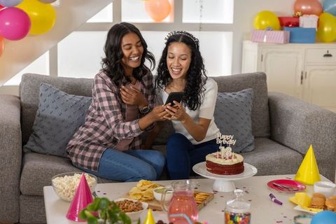 Smiling diverse sisters celebrating 18th birthday, sharing smartphone laughs on cozy couch