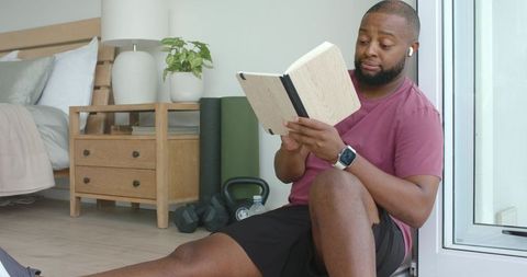 African american man reading notebook during home workout break with kettlebell and dumbbells