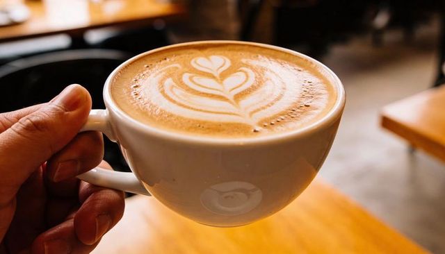 Hand holding latte cup showing leaf latte art over wooden table in cozy cafe