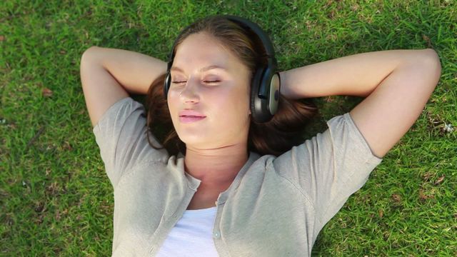 Young woman lying on green grass in a park wearing headphones, eyes closed, enjoying music. Ideal for concepts of relaxation, leisure, outdoor activities, wellness, connection with nature, and mindfulness.