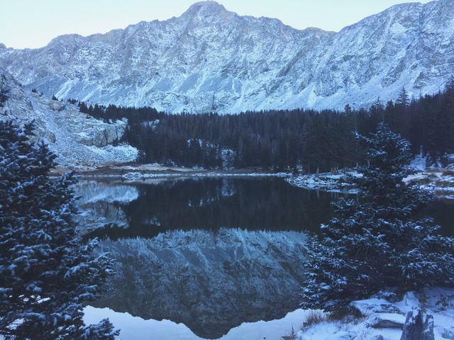 This tranquil scene captures a serene mountain lake surrounded by snow-covered pine trees and towering peaks. The reflection of the mountains in the calm water enhances the sense of peacefulness and natural beauty. This image is ideal for promoting travel destinations, outdoor activities, and winter adventures. It can also be used in backgrounds for inspirational themes, posters, or nature-related publications.