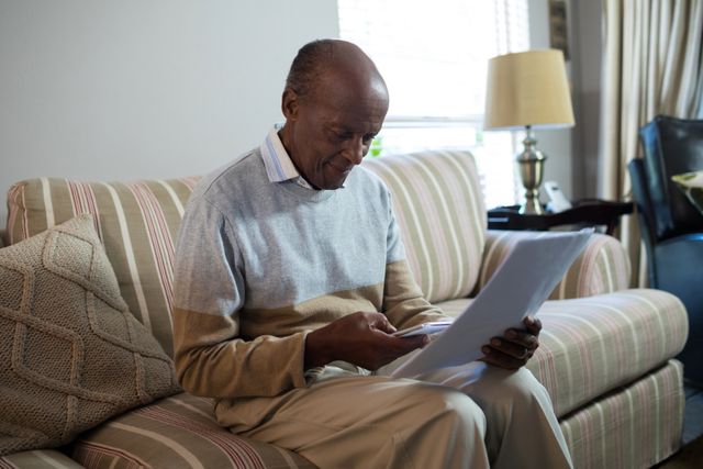 Senior man sitting on a striped sofa in a cozy living room, using a smartphone while holding a document. Ideal for themes related to elderly lifestyle, technology use among seniors, home comfort, and retirement activities.