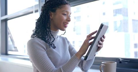 African American woman using tablet by window with coffee in modern minimalist workspace