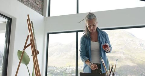 Mature female artist painting in sunlit loft studio holding glass and brush