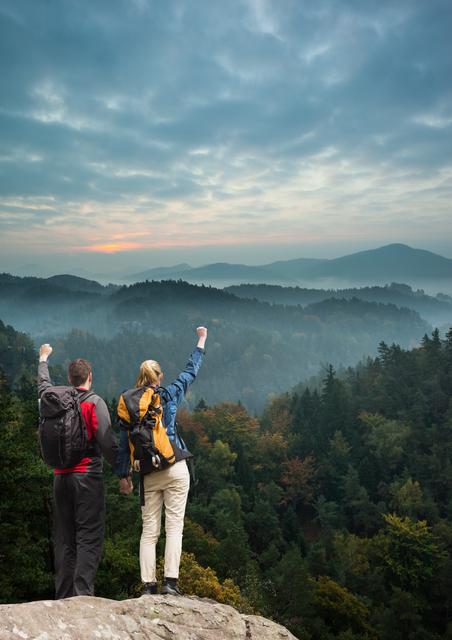 Couple standing on rock cliff raising arms in triumph overlooking forest and mountains at sunrise. Perfect for travel blogs, adventure and hiking promotions, outdoor activity advertisements, motivational content, and nature-themed websites.