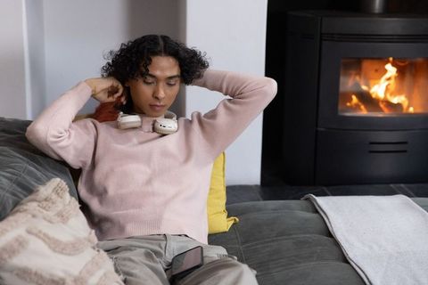 Nonbinary person relaxing on sofa by lit stove wearing pink sweater and headphones, cozy
