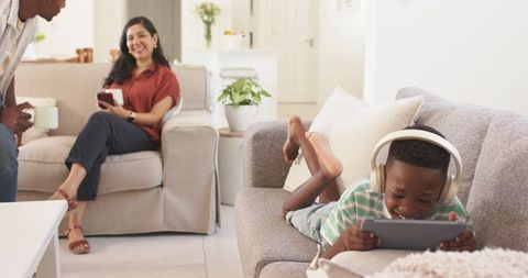Boy listening with headphones and playing tablet while family relaxing on modern sofa