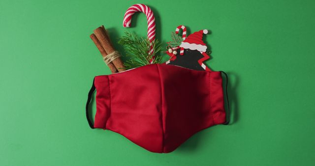 Depicts a festive red face mask with Christmas decorations such as candy cane, cinnamon sticks, and Santa ornament on green background. Perfect for illustrating holiday celebrations during pandemic, seasonal safety, Christmas-themed health campaigns, or festive holiday greetings.