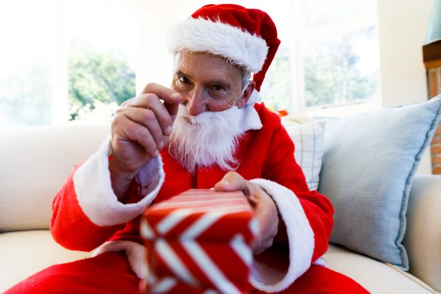 Front view of an old caucasian man wearing a santa clause costume holding out a christmas gift in front of him and pointing forward. he is sitting on the couch in the living room.