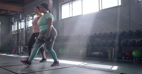 Two women performing walking lunges with dumbbells in sunlit gym