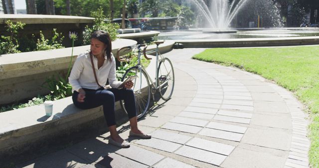 Woman is resting on a bench in a park on a sunny day with a bicycle leaning against the bench. She is holding a tablet, suggesting she is reading or browsing. The location features a fountain and greenery, providing a serene and relaxing environment. Ideal for concepts of relaxation, leisure, technology in everyday life, or outdoor activities.