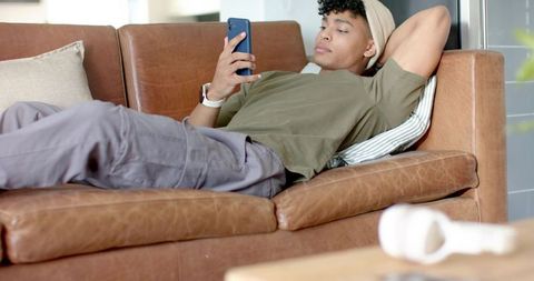 African american man reclining on brown leather sofa scrolling smartphone wearing beanie