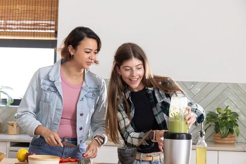 Female friends preparing green smoothie while cutting strawberries and using blender