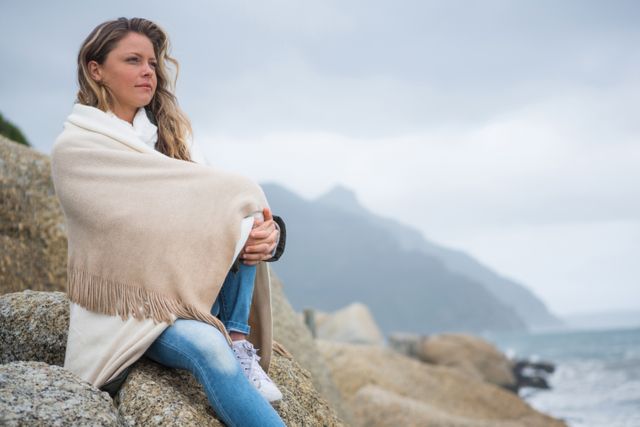 Woman wrapped in shawl sitting on rocks at beach, looking thoughtful. Ocean and mountains in background under cloudy sky. Ideal for themes of contemplation, solitude, nature, and peaceful moments. Suitable for use in wellness, travel, and lifestyle content.