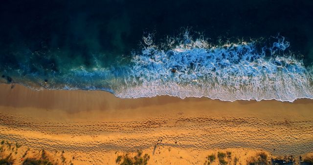 Aerial view of waves crashing onto a sandy beach, showcasing the contrast between the deep blue ocean and the golden sand. Ideal for travel and tourism promotions, summer vacation advertisements, nature and scenery websites, or environmental conservation materials.