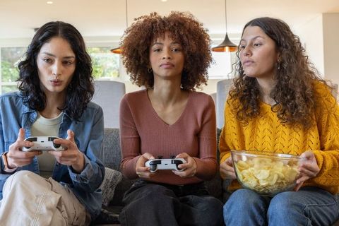 Three women playing video games on sofa sharing chips, relaxing in cozy living room