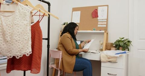 Woman reviewing fashion sketches and paperwork at small home studio desk with clothing rack