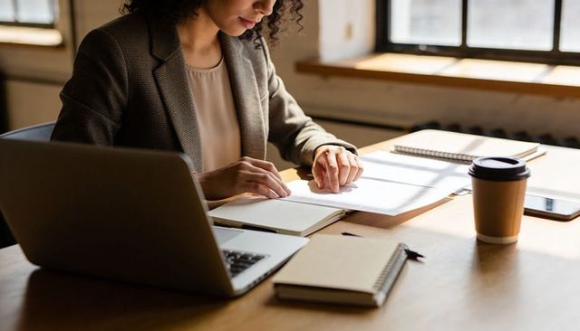 Focused professional reviewing paperwork at sunlit wooden desk with laptop and coffee