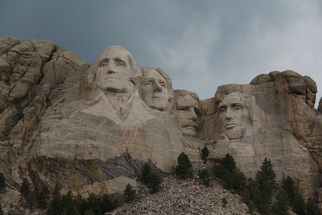Mount Rushmore shows four sculpture of U.S. presidents George Washington, Thomas Jefferson, Theodore Roosevelt, and Abraham Lincoln, carved into granite of Mount Rushmore. Overcast sky adds dramatic effect. Ideal for educational content, historical presentations, travel blogs, and patriotic themes.