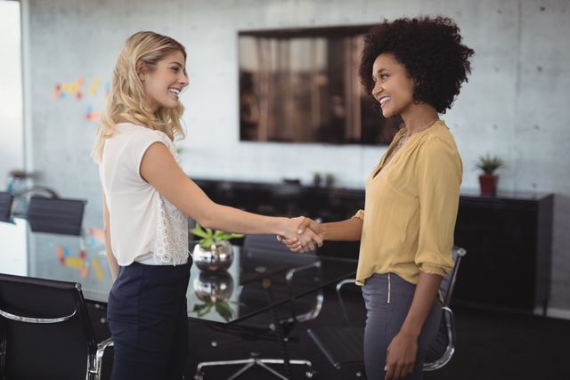 Two businesswomen are shaking hands in a modern office, indicating a successful meeting or agreement. This image can be used for themes related to business partnerships, professional collaboration, corporate success, and workplace diversity. Ideal for business websites, corporate presentations, and marketing materials.