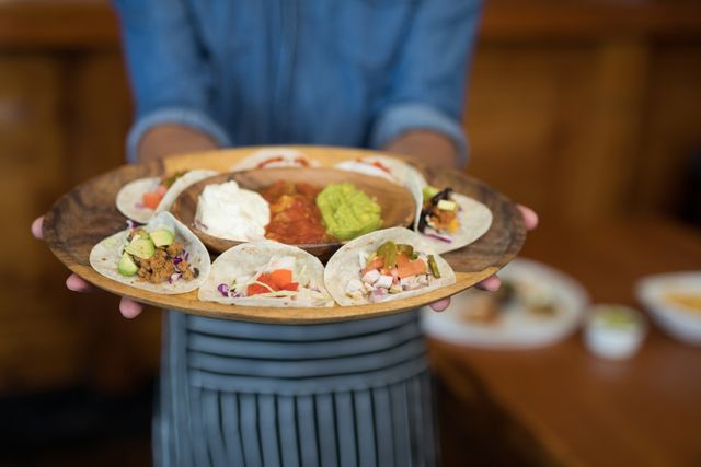 Waiter holding a wooden plate with an assortment of Mexican tacos and dips including guacamole, salsa, and sour cream. Ideal for use in content related to Mexican cuisine, restaurant dining, food service, hospitality industry, and culinary presentations.