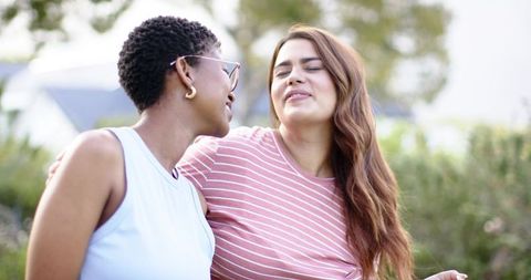 Two african american women laughing and embracing while standing close in sunlit garden